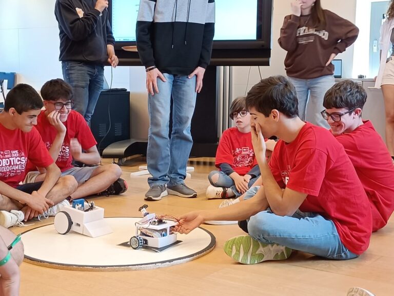 Alumnes d’R7 participant a la competició de robots de sumo Gitech, asseguts al voltant d’un taulell mentre dos robots es preparen per competir.
